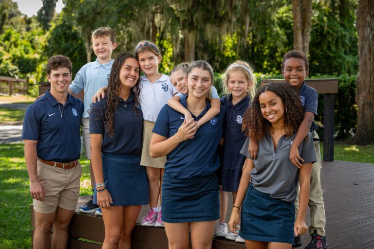 A group of young students and older students in uniform pose together outdoors on a wooden platform with trees in the background.