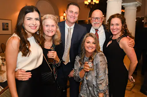 A group of six adults, dressed in formal attire, pose and smile for the camera at an indoor event or gathering.