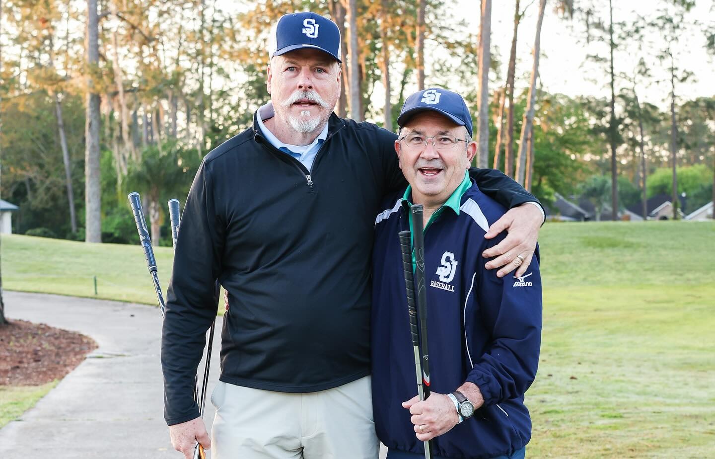 Two men wearing SJ hats and jackets stand side by side on a golf course, each holding a golf club, with trees and grass in the background.