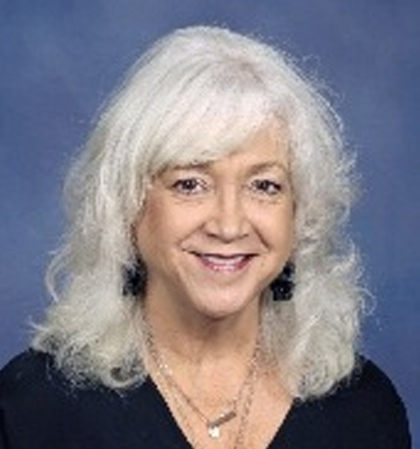 Woman with shoulder-length white hair wearing a black top and layered necklaces, smiling at the camera against a blue background.